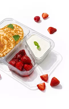 Three-compartment clear container holding pancakes with mint, yogurt in the top right, and diced strawberries in the bottom left; scattered whole and halved strawberries on a white surface.
