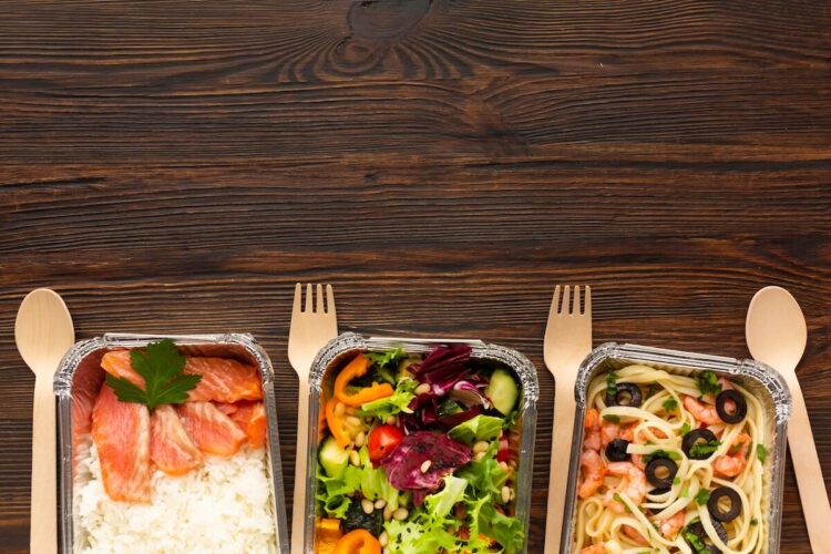 jak-podgrzewac-catering-dietetyczny (2) | Catering dietetyczny | Dieta pudełkowa | Pomelo | Catering dietetyczny | Dieta pudełkowa | Pomelo Three meal containers on a dark wooden table: salmon with rice, a colorful veggie salad, and shrimp pasta with olives.