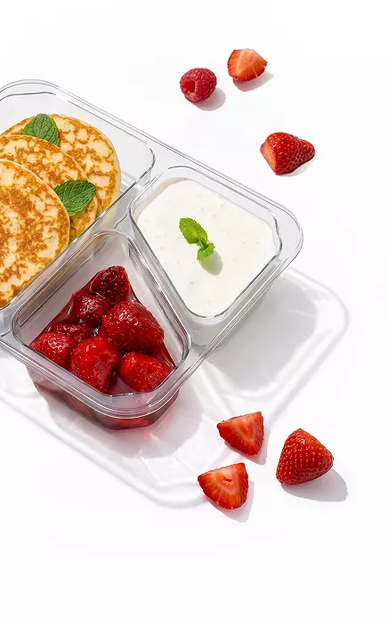 Three-compartment clear tray with mini pancakes and mint leaves, a yogurt dip topped with mint, and a bowl of strawberries; scattered berries nearby on a white surface