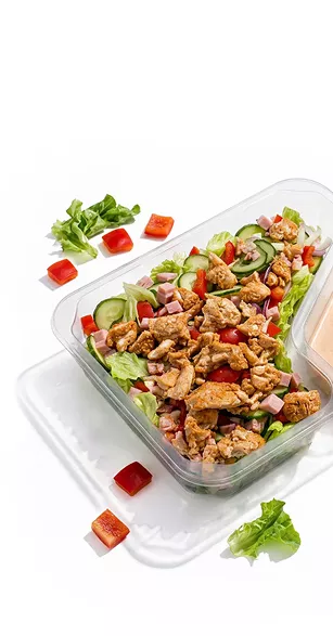 Glass meal container holding a chicken salad with lettuce, cucumber, tomatoes, red onion, and diced peppers.
