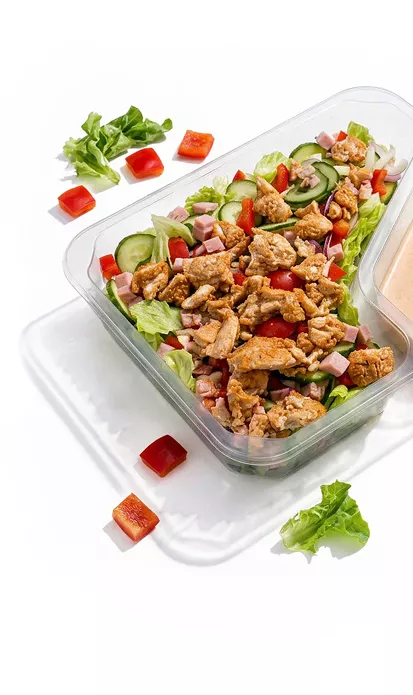 Chicken salad in a clear glass container with lettuce, cucumber, tomatoes and ham cubes, plus dressing on the side