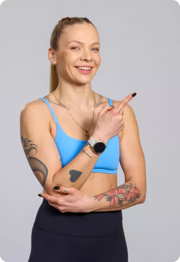 Smiling athletic woman in a blue sports bra points to the right; has tattoos on her arms and wears a watch against a gray background.
