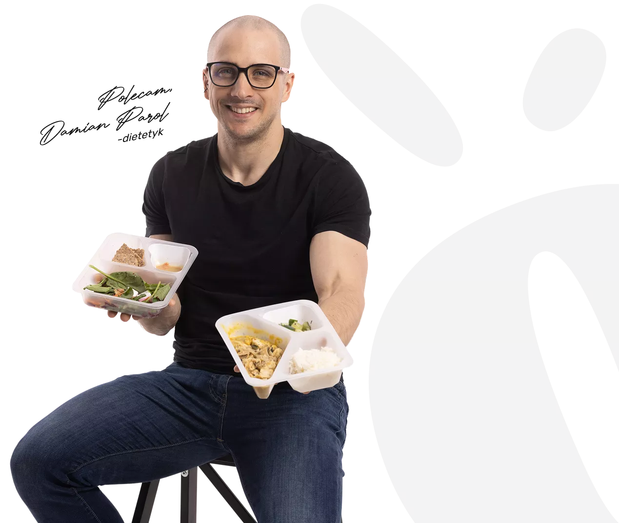 Smiling man with glasses holding two meal-prep boxes of salad and wrap, seated against a dark background.