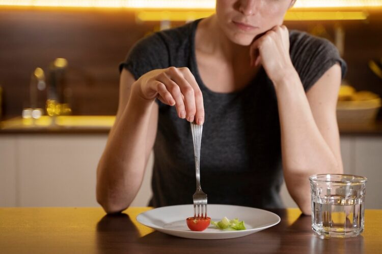 brak-apetytu-w-upaly (1) | Catering dietetyczny | Dieta pudełkowa | Pomelo | Catering dietetyczny | Dieta pudełkowa | Pomelo Person sitting at a table using a fork to pick up a cherry tomato from a plate, with a thoughtful pose and a glass of water nearby.