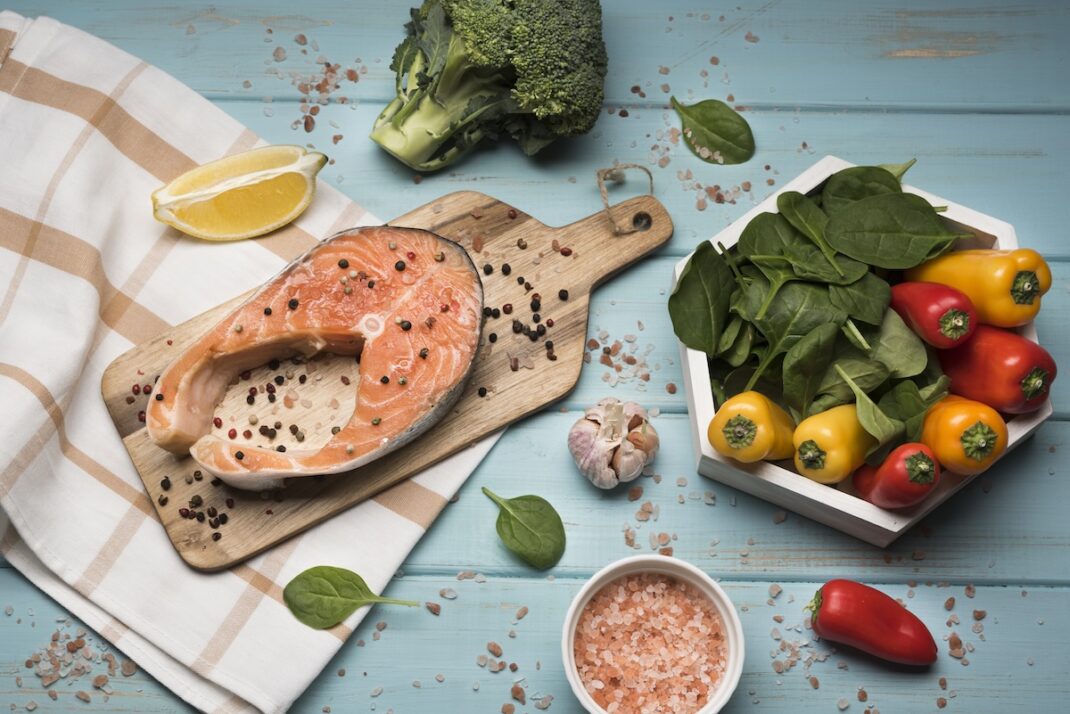 Salmon steak on a wooden board with peppercorns, a lemon wedge, garlic, broccoli, spinach, and a colorful assortment of peppers on a blue wooden table.