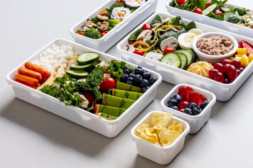 White meal-prep boxes brimming with fresh veggies, fruit, and salads—rice with carrots and cucumber, avocado slices, kale, and berries visible in each container.