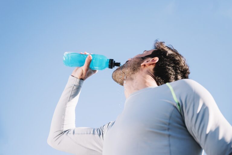 elektrolity | Catering dietetyczny | Dieta pudełkowa | Pomelo | Catering dietetyczny | Dieta pudełkowa | Pomelo Man in a light athletic shirt tilts his head back to drink from a blue water bottle against a clear blue sky.
