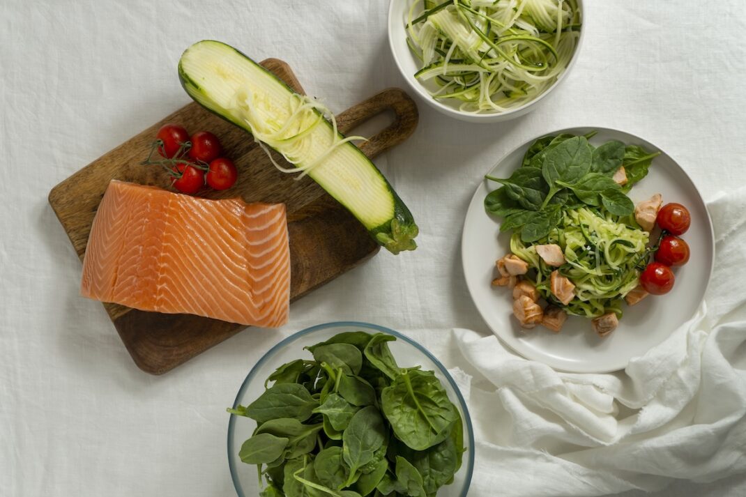 Salmon fillet on a wooden cutting board with zucchini ribbons and cherry tomatoes, plus bowls of greens and zucchini noodles nearby.