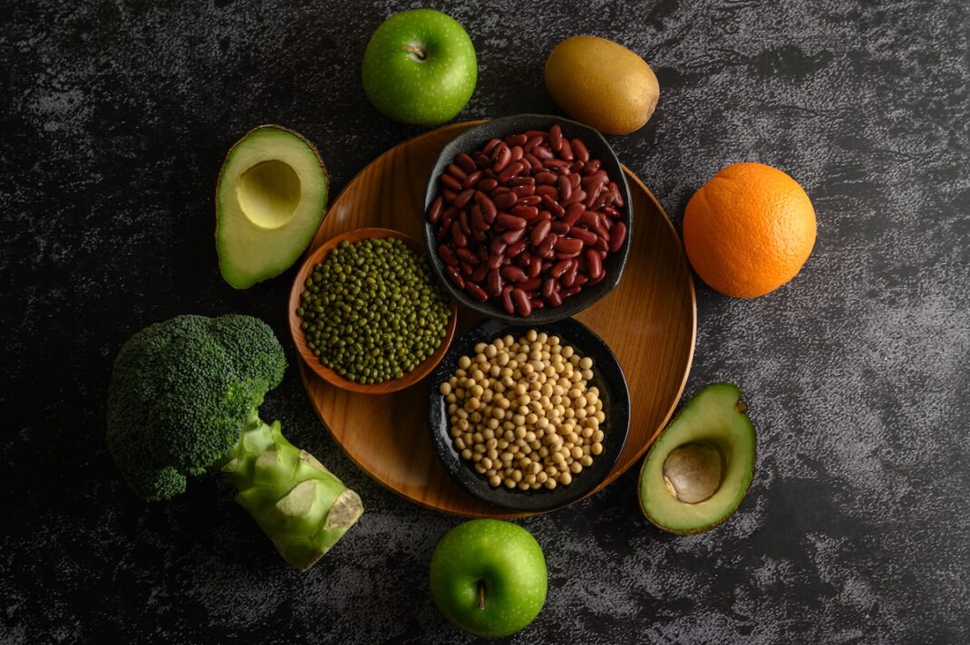 Top-down view of a wooden board with bowls of red beans, green lentils, and chickpeas, surrounded by avocado halves, broccoli, apples, kiwis, and oranges on a dark textured surface.