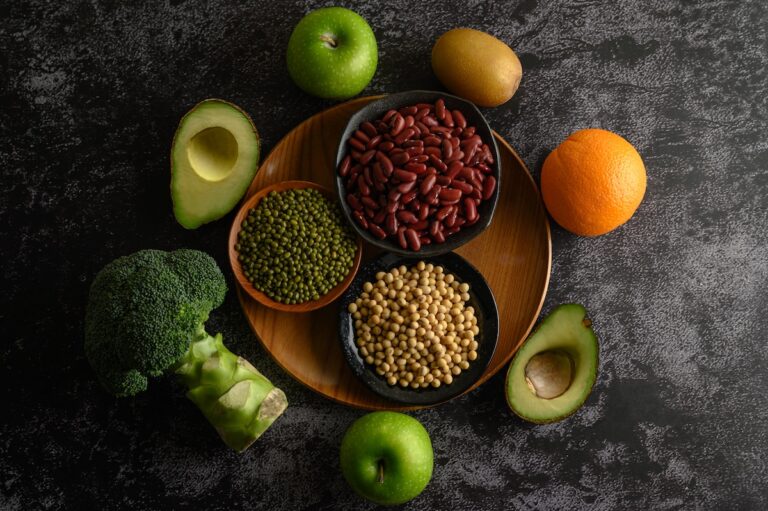 roslinne-zrodla-bialka | Catering dietetyczny | Dieta pudełkowa | Pomelo | Catering dietetyczny | Dieta pudełkowa | Pomelo Top-down view of a wooden board with bowls of red beans, green lentils, and chickpeas, surrounded by avocado halves, broccoli, apples, kiwis, and oranges on a dark textured surface.