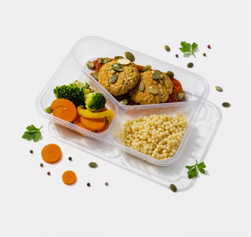 Three-compartment plastic meal tray holding broccoli and carrot medley with yellow pepper, tomato sauce topped with seed-covered cookies, and pearl couscous; parsley and seeds scattered nearby for garnish.