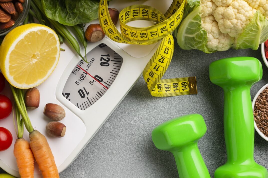 Healthy eating and fitness theme: scale, measuring tape, vegetables, lemon half, nuts, and bright green dumbbells on a workout surface.