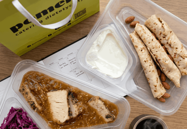Meal prep containers on a wooden table: sliced chicken in sauce, yogurt dip, flatbreads with almonds, and purple cabbage side.